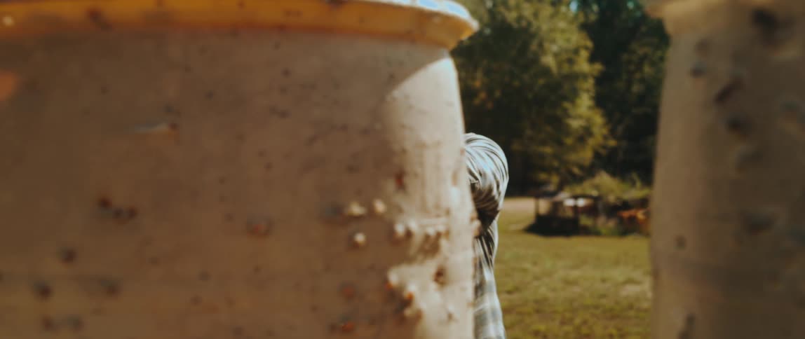 A man in a flannel shirt and ear protection practices tactical shooting at an outdoor range moving between large plastic barrels while aiming and firing a handgun equipped with a red dot sight toward the camera