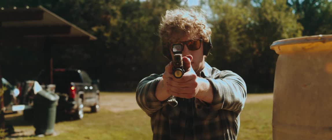 A man wearing sunglasses and ear protection steps out from behind a yellow barrel while aiming a handgun directly forward he maintains a steady grip on the firearm with both hands as he moves into a clear view against an outdoor background