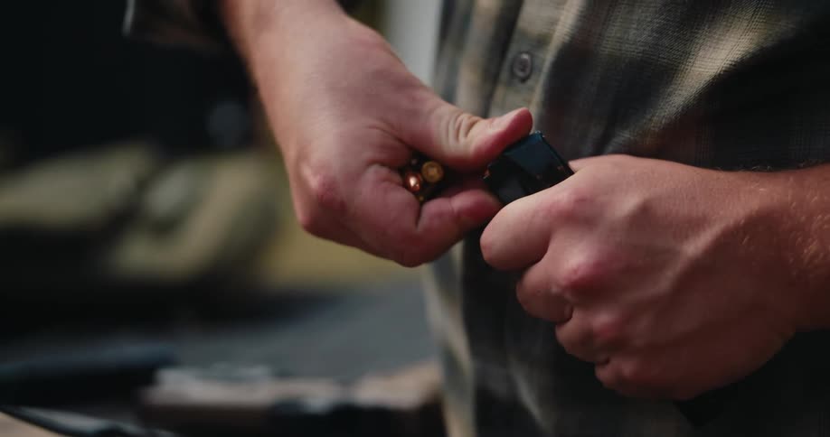 A person wearing a plaid shirt holds several goldcolored rounds in one hand while using the other to load them into a black pistol magazine they carefully press each cartridge down into the magazine repeating the process with multiple rounds