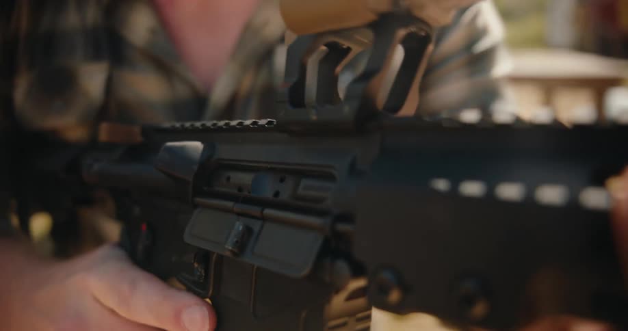A rifle is being fired outdoors by a man with spent casings and debris ejecting from the weapon as it discharges