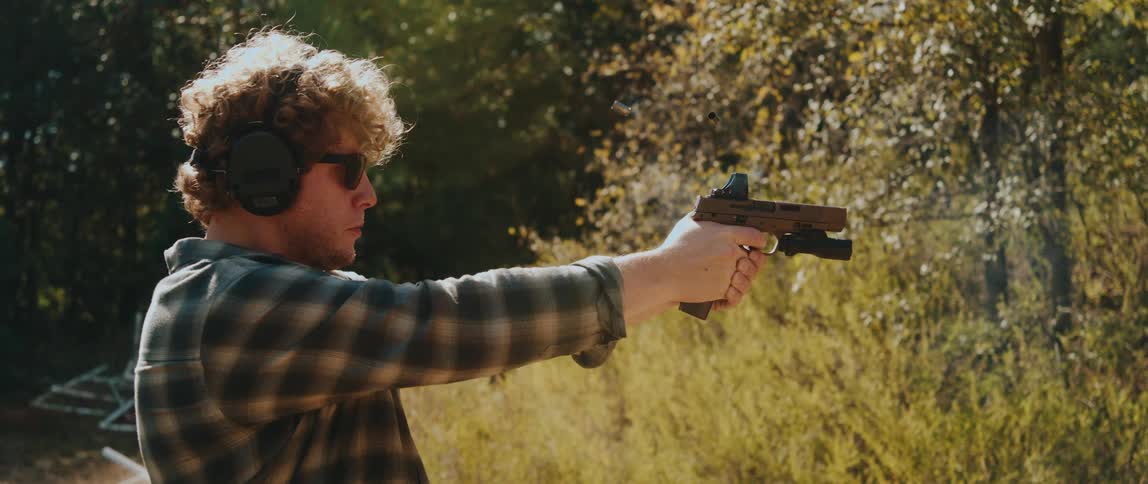 Man wears ear protection and safety glasses while firing a handgun in a wooded outdoor area