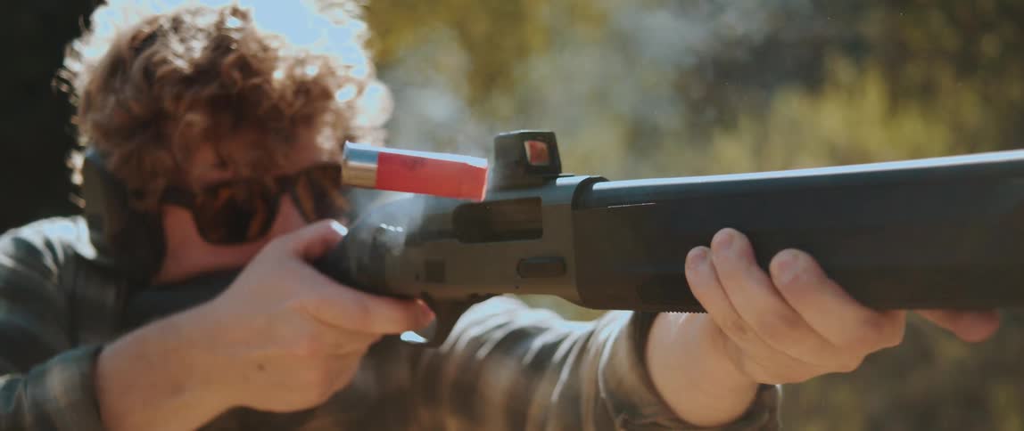 A man practices with his shotgun in the forest as a red shell casing arcs through the sunlight