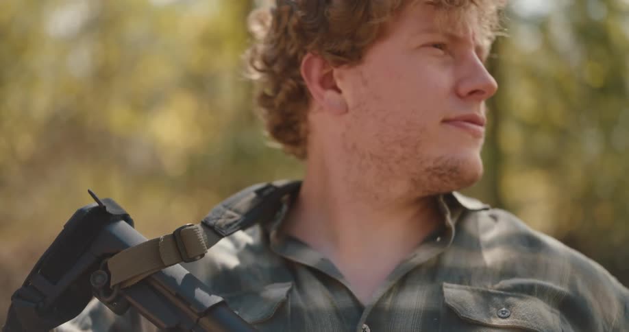A curlyhaired man in a plaid shirt holds a rifle while standing outdoors looking from the side toward the camera and breaking into a slight smile as he adjusts his grip on the firearm