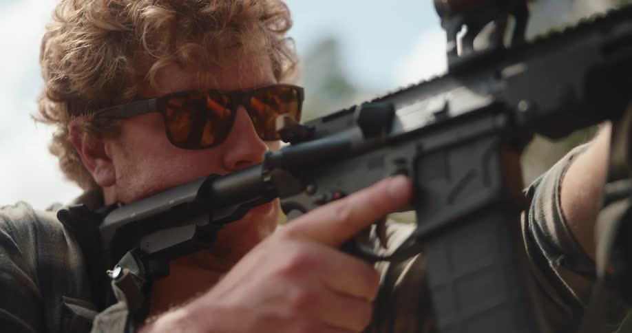 A man in a plaid shirt inspects and then aims a rifle looking down its sights