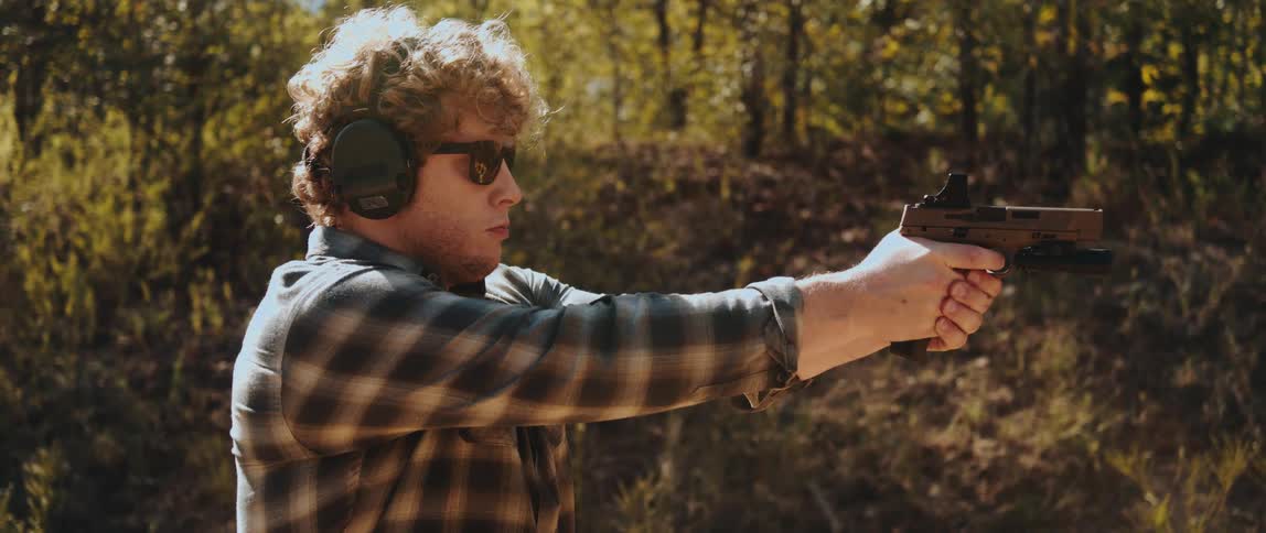 A man fires a handgun at an outdoor shooting range surrounded by trees