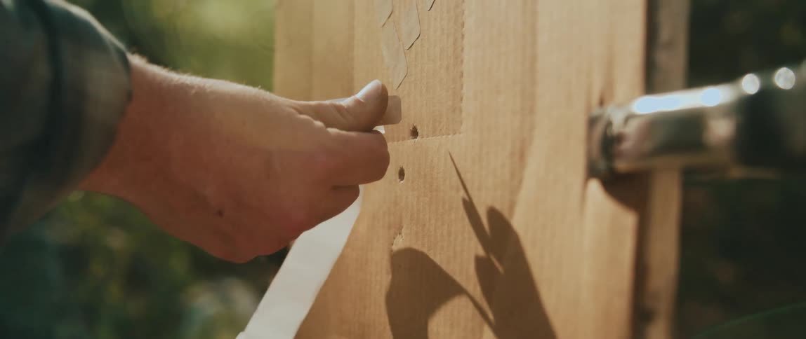 A closeup shows a person holding a small handheld level against a pale wooden surface with sunlight highlighting the wood grain tool edges and the alignment between the level and the vertical board