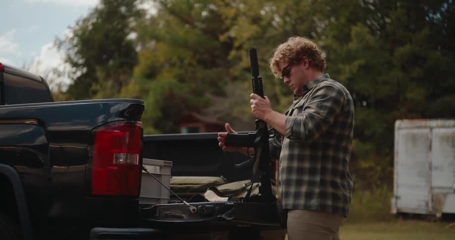 In this short clip a man with curly hair stands at the tailgate of a black pickup truck and prepares a rifle he carefully inserts a magazine into the firearm while standing in an outdoor wooded setting