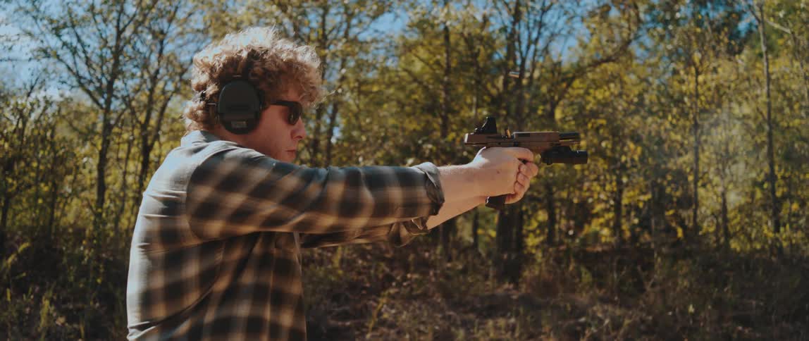 A young man wearing headphones firing a weapon at an outdoor shooting range in the woods at midday