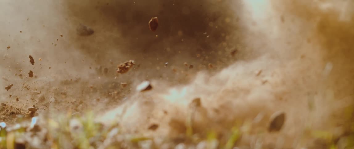 A groundlevel closeup captures dirt dust and small rocks erupting into the air above grass