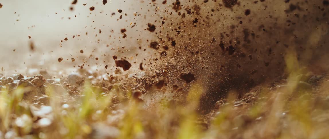 Dirt and rocks explode violently from the ground in a closeup slowmotion shota cloud of dust and debris hangs in the air above a sunlit grassy landscape