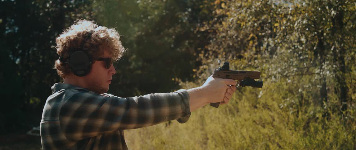 A man wearing ear protection and sunglasses fires a tan semiautomatic pistol at an outdoor range with smoke and a spent shell casing ejecting from the firearm as the slide cycles back during the shot
