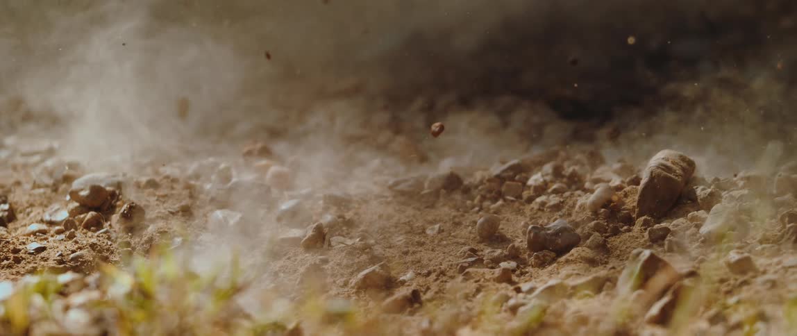 A groundlevel explosion blasts dirt rocks and dust into the air with debris raining back down onto the dry soil and patches of grass in slow motion as the smoke clears