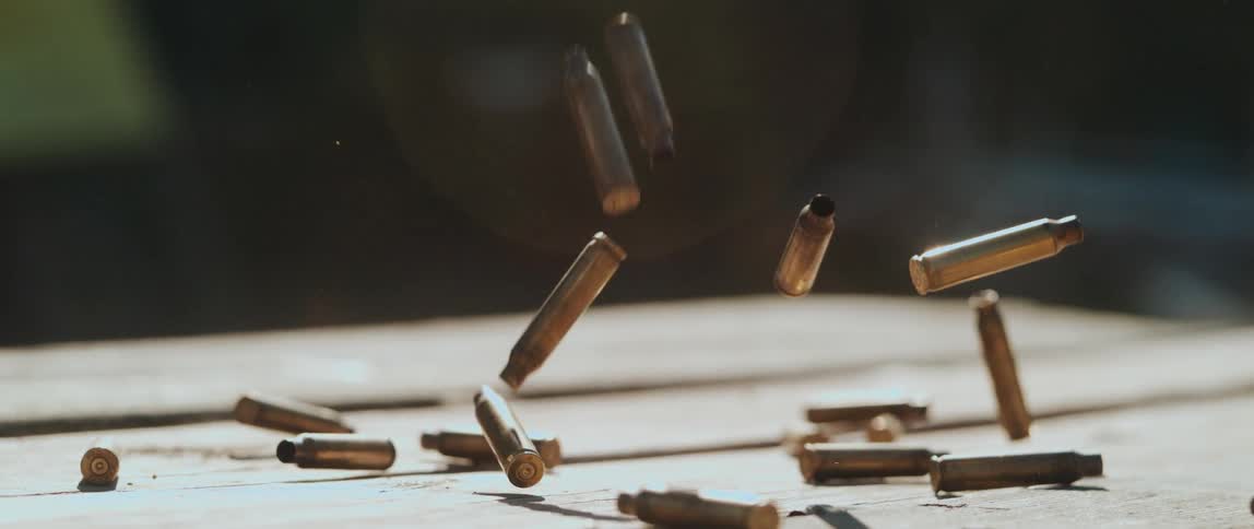 In slow motion empty brass bullet casings cascade down bouncing and scattering across a sunlit wooden surface