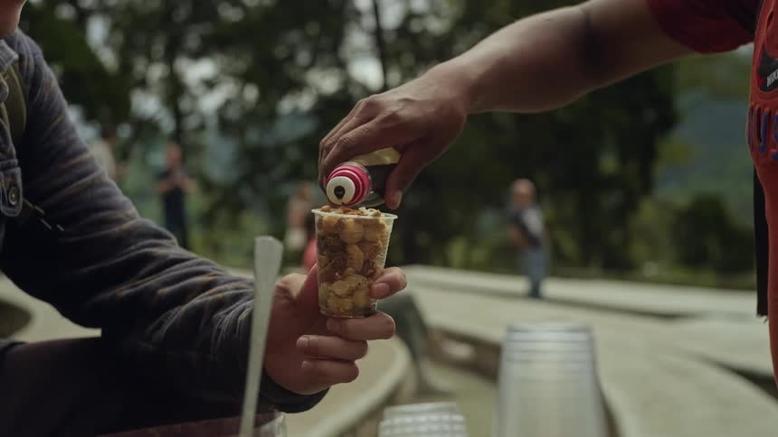 A person pours a dark syrup from a small bottle into a clear plastic cup filled with chopped fruit and nuts held by another person in an outdoor park setting with trees and blurred figures in the background