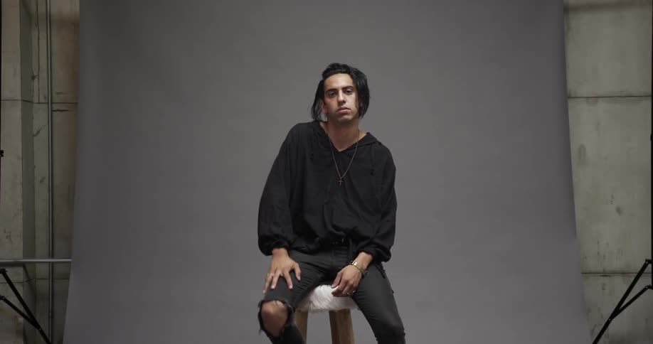 A greasy haired man sits on a stool