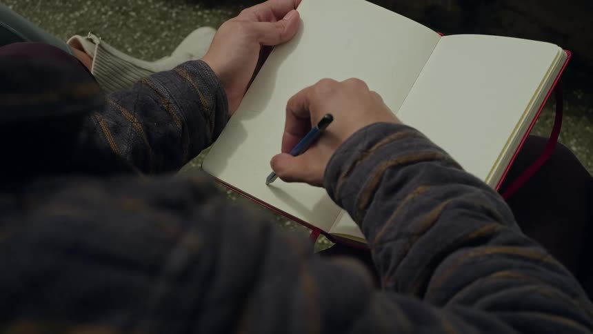 A closeup view from over the shoulder shows a persons hands using a mechanical pencil to draw thin vertical lines onto the blank white page of a small redbound notebook