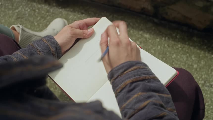 A manwearing a striped jacket and purple pants sits outdoors while using a blue pencil to sketch a cross symbol into a small redbound notebook held in his lap