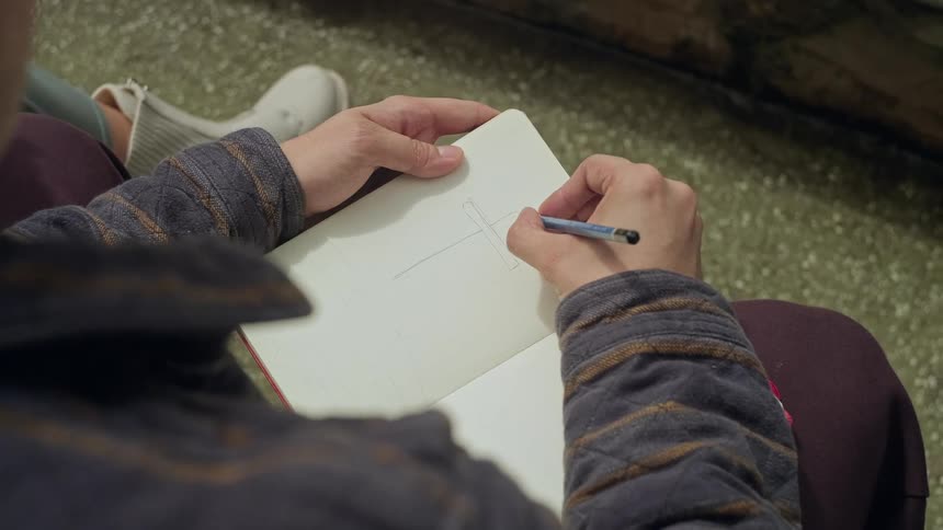 A topdown closeup shows a person sketching with a pencil in an open notebook resting on their lap with both hands visible and the surrounding ground and shoes partially in frame