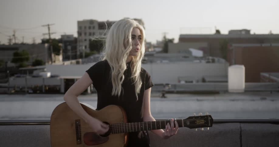 A woman with blonde hair plays guitar on a roof