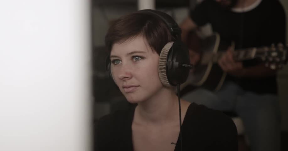 A woman wears headphones as another plays guitar