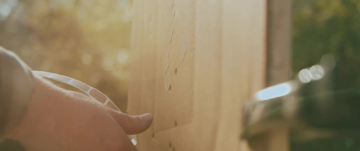 A closeup shows a hand peeling or pressing a strip of tape against a vertical wooden surface with visible holes and grain while sunlight illuminates the wood and a metal fixture appears at the edge of the frame