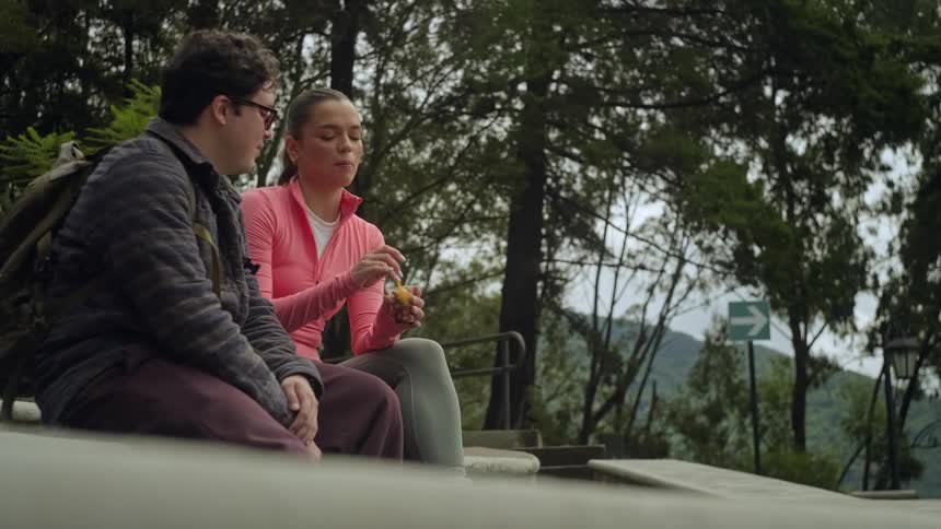A man with glasses and a backpack sits on a stone ledge next to a woman in a pink shirt as they eat snacks together in a park with tall trees