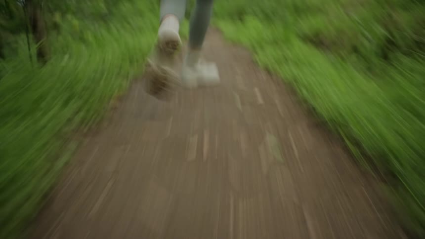 A lowangle shot shows a person running along front and backwards on a narrow dirt path with legs and shoes in motion as greenery blurs along both sides of the trail
