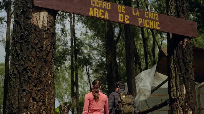 Two people walk beneath a wooden sign reading cerro de la cruz area de picnic entering a forested picnic area framed by tall trees with a tarpcovered structure visible to the side