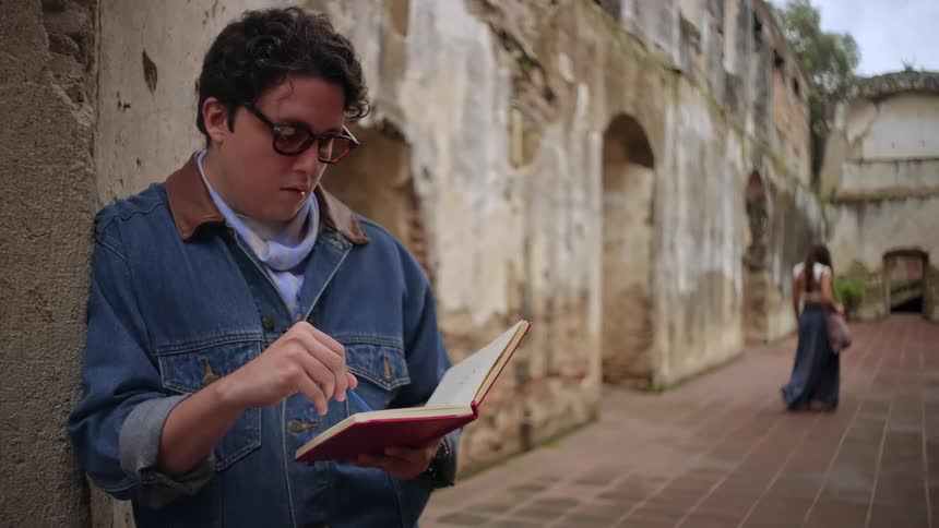 The man leans against the stone wall while writing in his notebook