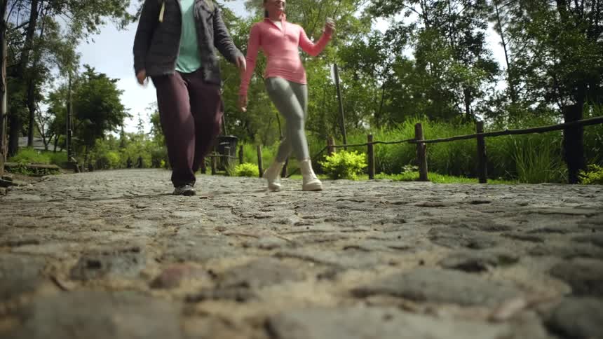 A man and a woman walk toward the camera on a wide cobblestone path lined with a wooden fence and dense green trees