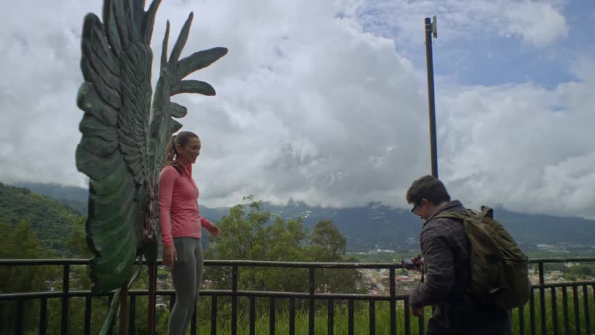 A woman in a pink shirt stands in front of a large angel wing sculpture smiling and gesturing as a man takes her photo with a camera
