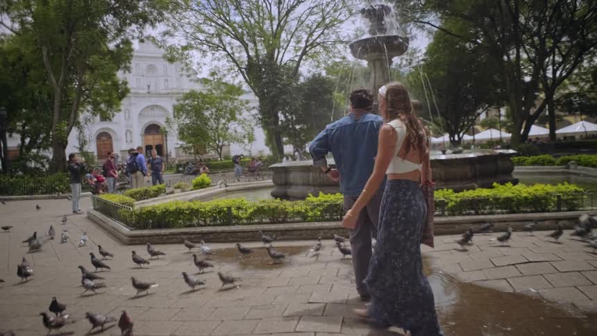 A couple walks through a park filled with pigeons the man lifting the womans hand as they stop at a fountain the woman brushing her hair back from her face as they stand together