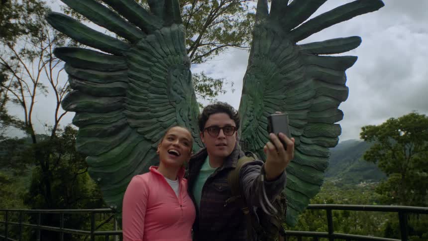 A young man in glasses and a woman in pink pose for a selfie in front of a large green wing sculpture at a scenic mountain overlook