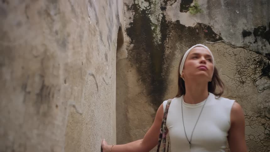 A woman wearing a white headband and crop top walks through a narrow stone corridor in an ancient ruin looking up at the high weathered walls as she explores the textured passage
