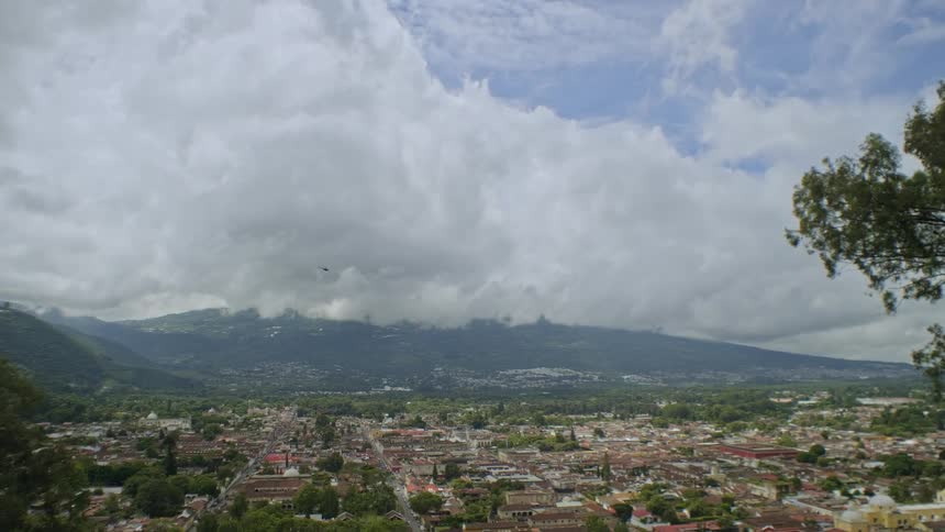 A wide panoramic view shows a dense town of lowrise buildings spread across a green valley with mountains rising in the distance beneath thick layered clouds and patches of blue sky