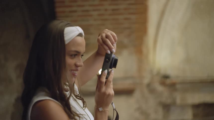 A woman with a white headband holds a small camera up near her face to take a selfie adjusting it with both hands while standing beside a brick and stone architectural background