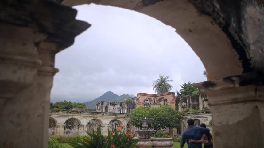 A couple stands with their arms around each other gazing out at the sprawling stone ruins of a colonialera estatethe scene is framed by a crumbling archway revealing a lush courtyard with a fountain and misty green mountains in the distance
