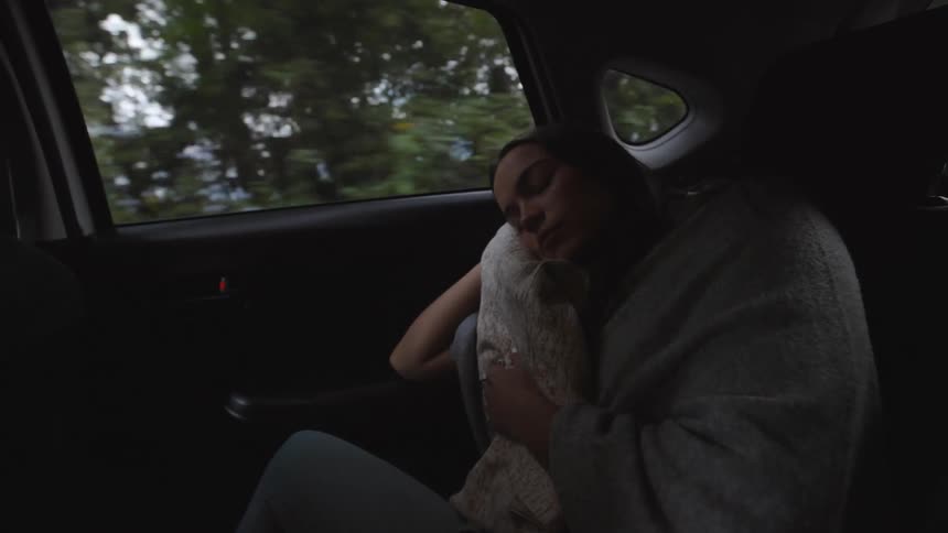 A woman sleeps peacefully in the backseat of a moving car holding a blanket as scenery passes by the window