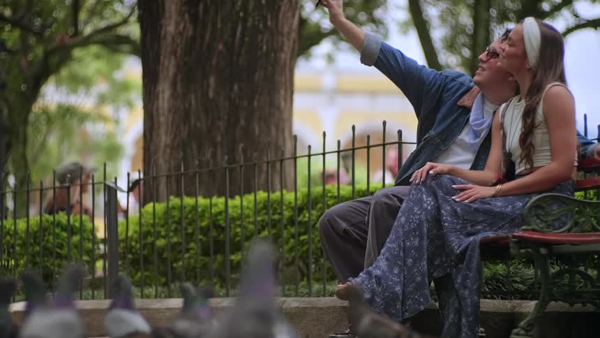 A man and woman sit on a park bench smiling as he takes a selfie of them with his smartphone