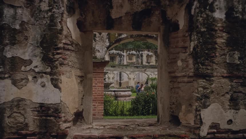 A couple walking hand in hand through a garden surrounded by ancient ruined buildings at midday
