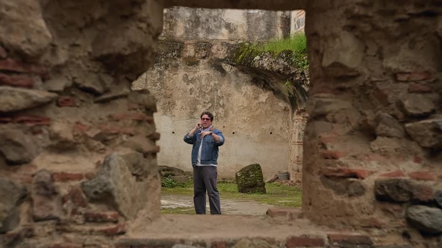 Viewed through a weathered stone window a man casually talks on his phone while standing in the mosscovered courtyard of ancient ruins