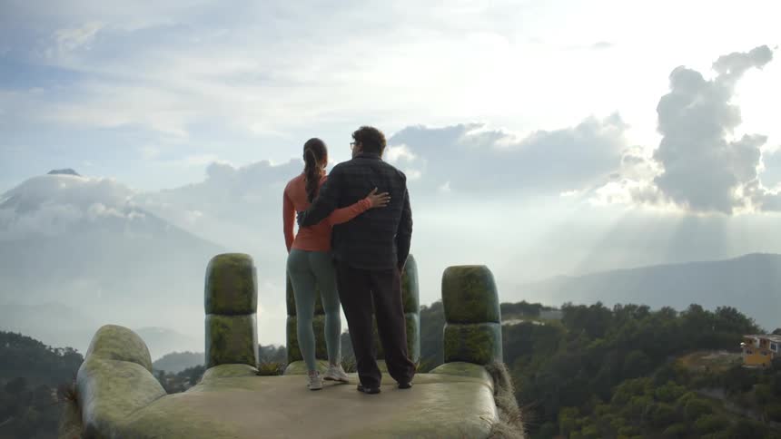 A couple standing on the statue palm at hobbitenango ecopark hugging as they look out over the vast landscape on a cloudy day