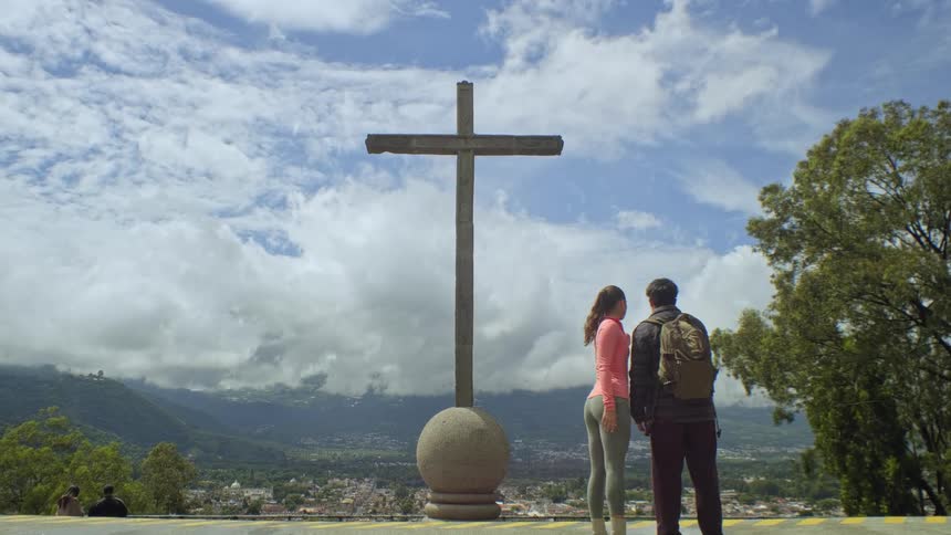 A couple in hiking gear stands handinhand at a scenic overlook looking past a large stone cross toward a valley town and misty mountains under a bright sky