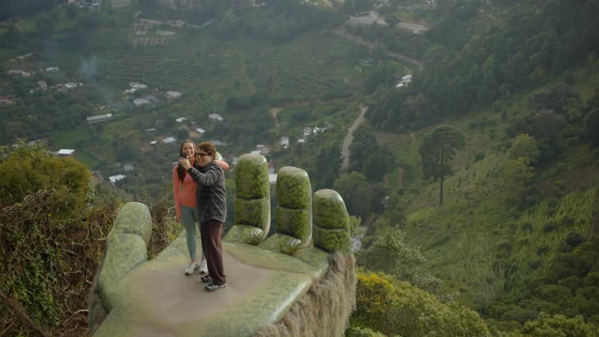 A woman in an orange shirt and a man stand on a large stone hand sculpture overlooking a lush valley where they pose together to take a selfie against the backdrop of a misty mountain village and dense green forests