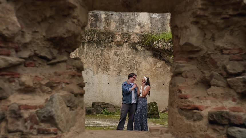 A view through a stone window frame shows a man in a denim jacket and a woman in a long skirt standing together and talking in a courtyard with ancient ruins