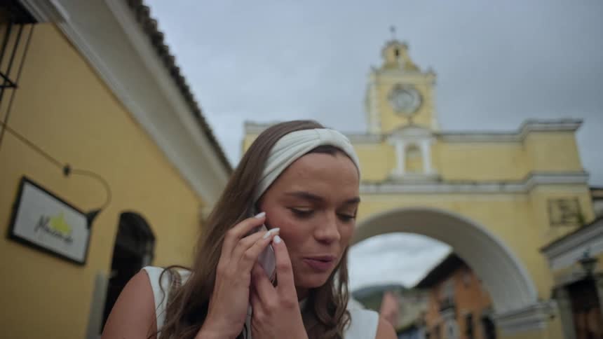 The video shows a woman with a white headband talking on a smartphone in a historical city centershe is standing in front of a prominent yellow archway with a clock tower likely the santa catalina arch in guatemala