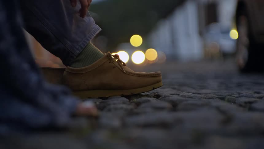 A person standing on the side of the road at evening time wearing brown shoes as cars drive by