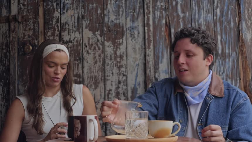 Two people seated at a wooden table with mugs and a glass pitcher as one pours liquid in a mug framed against a weathered wooden door backdrop