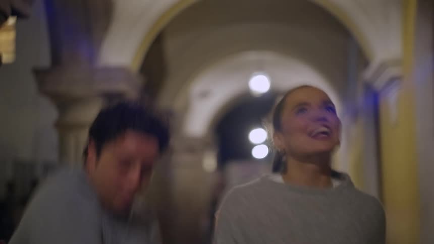 A man and a woman walk toward the camera through a stone archway at night laughing and talking and the scene is lit by warm overhead lights and distant traffic