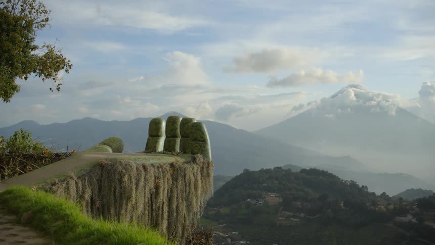 A large stone hand sculpture sits on the edge of a mountain cliff overlooking a lush valley with rolling hills and a distant volcano under a cloudy blue sky